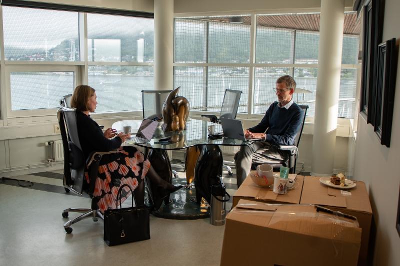 Ei kvinne og ein mann sitt på eit ganske tomt kontor, med kvar sin datamaskin ved eit glassbord. Ein rimeleg stor skulptur står mellom dei.